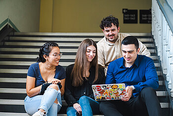 Gruppe von Studierenden sitzt auf einer Treppe und schaut gemeinsam auf einen Laptop
